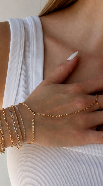 Close-up of a hand resting on the collarbone wearing layered gold and silver chain bracelets and a delicate chain ring over a white ribbed tank top — minimalist jewelry, summer fashion detail