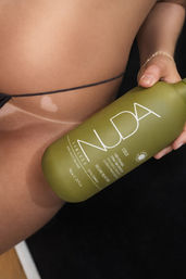 Close-up of a bikini-clad midriff with tan lines and a small heart-shaped lighter patch, hand holding an olive-green sunless tanning spray bottle for a beach-ready glow