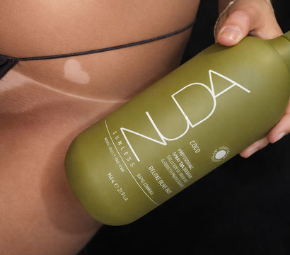 Close-up of a bikini-clad midriff with tan lines and a small heart-shaped lighter patch, hand holding an olive-green sunless tanning spray bottle for a beach-ready glow