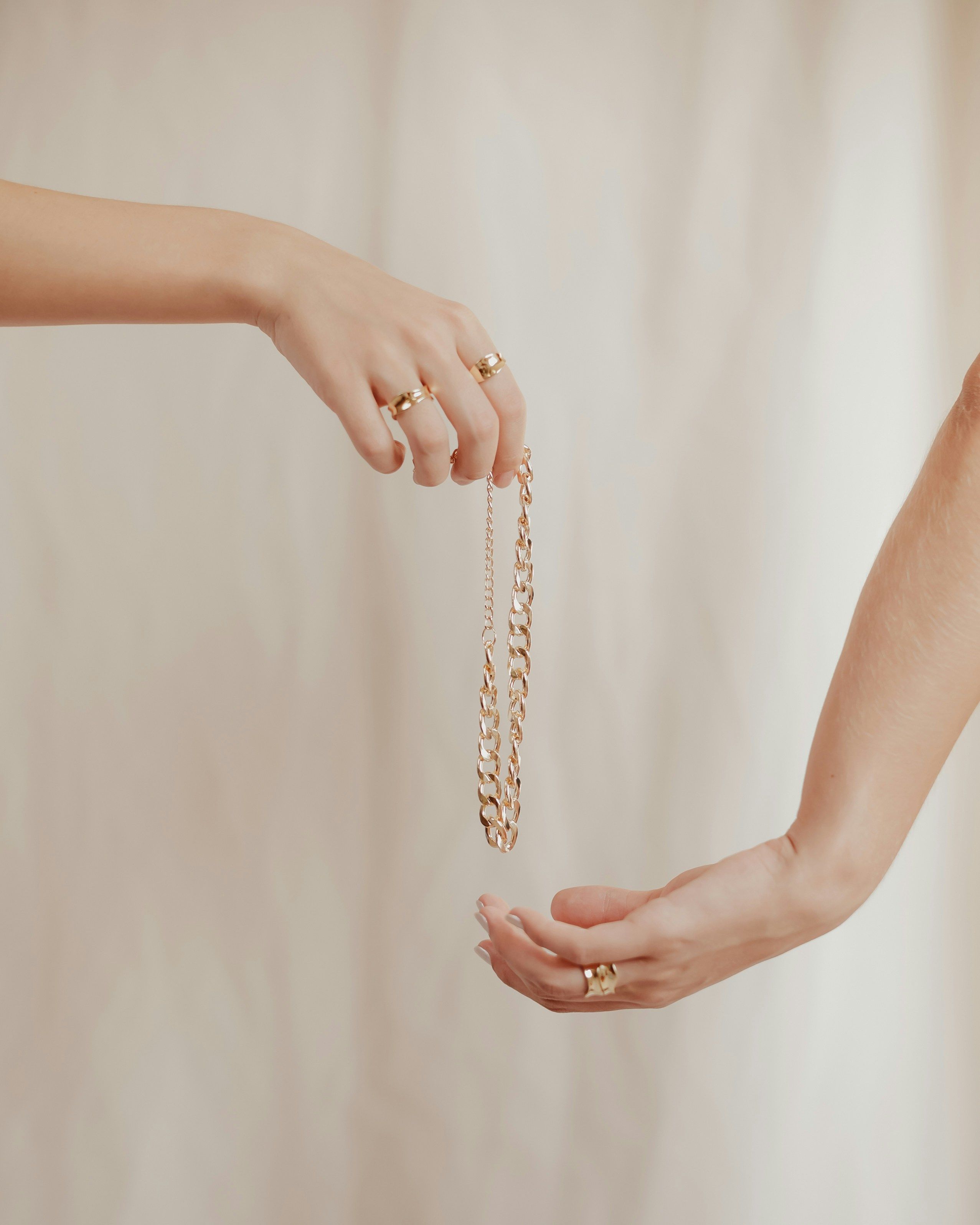Close-up of two hands passing a chunky gold chain necklace, both wearing gold rings, against a soft beige fabric backdrop.