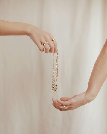 Close-up of two hands passing a chunky gold chain necklace, both wearing gold rings, against a soft beige fabric backdrop.