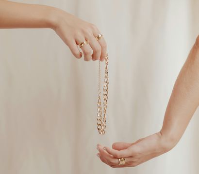 Close-up of two hands passing a chunky gold chain necklace, both wearing gold rings, against a soft beige fabric backdrop.