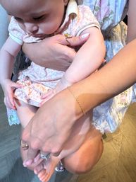 Close-up of a baby in a pink patterned dress cradled in an adult's arms, showing hands with gold rings and a delicate bracelet, floral fabric and hardwood floor background.