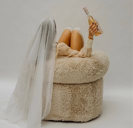 Veiled bride lounging on a plush round chair in a neutral studio, holding a champagne bottle with lace garter‑clad legs — playful bridal boudoir shot
