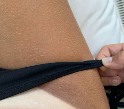 Close-up of sun-tanned skin showing a sharp white bikini tan line as a hand pulls a black swimsuit strap on a white lounge cushion — beachwear sunbathing detail.