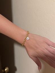 Close-up of a hand and wrist wearing a gold chain bracelet with a round white charm engraved with the letter H, set against a neutral beige indoor wall.