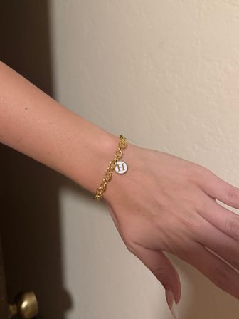 Close-up of a hand and wrist wearing a gold chain bracelet with a round white charm engraved with the letter H, set against a neutral beige indoor wall.