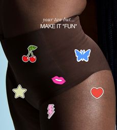 Close-up of a torso in brown high-waist swim bottoms decorated with colorful sticker graphics (cherries, blue butterfly, pink lips, star, lightning bolt, heart) and overlaid text reading 'your tan but... MAKE IT *FUN*'.