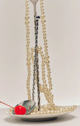 Still-life of creamy pearl necklaces draped over a twisted metal stand, pearls spilling onto a white plate with a reflective spoon holding a bright red maraschino cherry against a soft beige background.