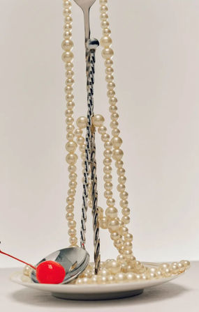 Still-life of creamy pearl necklaces draped over a twisted metal stand, pearls spilling onto a white plate with a reflective spoon holding a bright red maraschino cherry against a soft beige background.