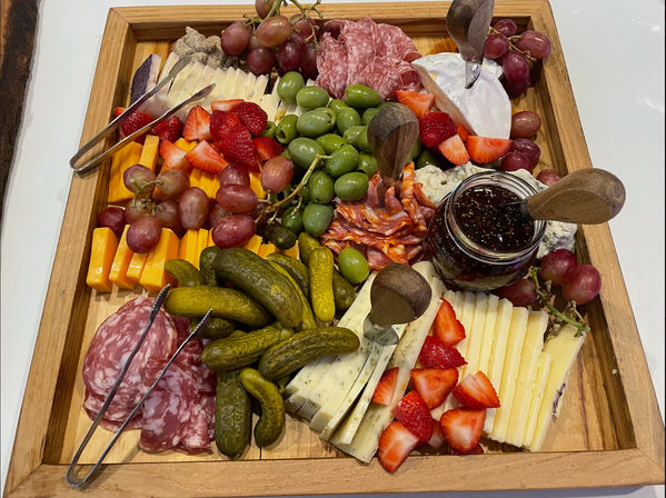 Vibrant square wooden charcuterie board with sliced cheeses, salami and pepperoni, green olives, cornichons, strawberries, red grapes and a jar of dark jam — party-ready