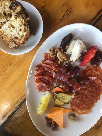 Charcuterie plate on a wooden table with sliced cured meats, cluster of red grapes, cheddar and blue cheeses, pickles, nuts, strawberries, jam and a side bowl of artisanal sourdough