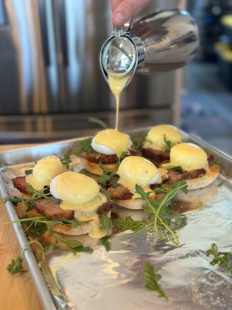 Close-up brunch shot of eggs Benedict on toasted English muffins with seared ham and arugula, warm hollandaise sauce being poured from a stainless pitcher.