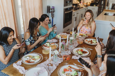 Group of friends enjoying a home brunch in a modern kitchen, seated around a decorated dining table and snapping photos of plated brunch dishes and cocktails.