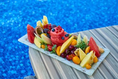 Poolside tropical fruit platter with a watermelon‑sorbet tower topped with blueberries and red currants, surrounded by mango, pineapple, cherries, strawberries and passionfruit on a striped lounge chair by a bright blue pool