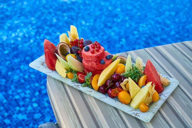 Poolside tropical fruit platter with a watermelon‑sorbet tower topped with blueberries and red currants, surrounded by mango, pineapple, cherries, strawberries and passionfruit on a striped lounge chair by a bright blue pool