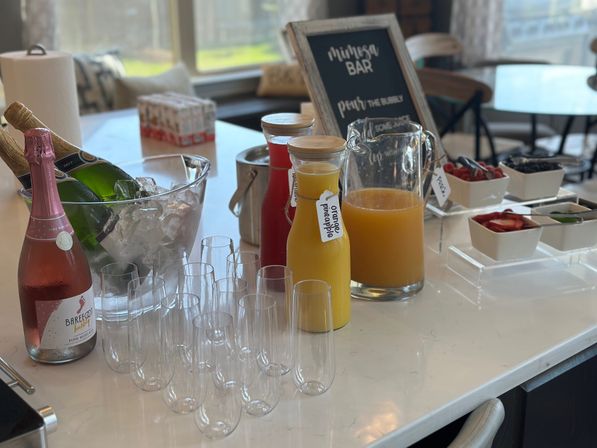 Brunch mimosa bar on a bright kitchen island with sparkling wine on ice, carafes of orange and pineapple juice, rows of clear champagne flutes and bowls of fresh fruit garnishes.