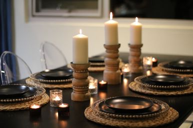 Warm candlelit dining table with three wooden pillar candle holders, votive candles, black plates on woven rattan placemats, cozy modern table setting