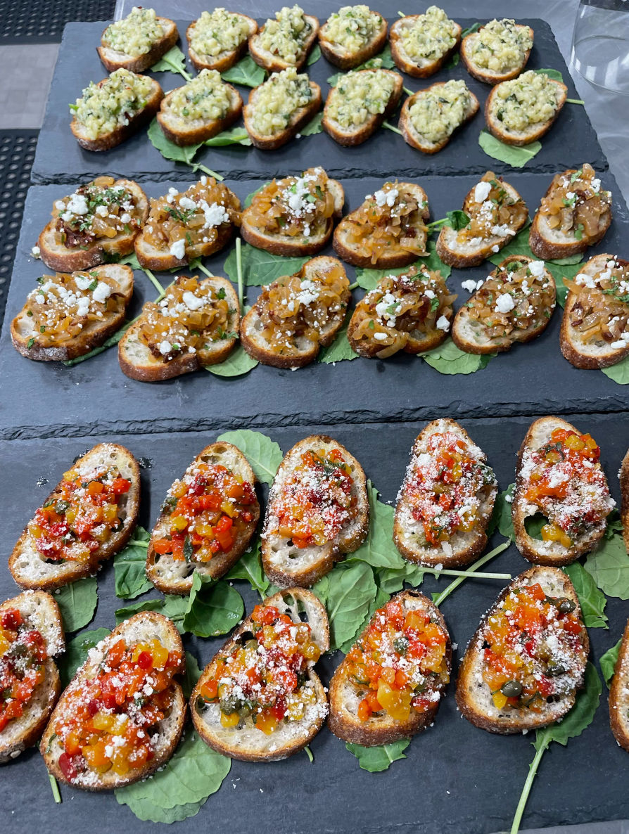 Gourmet appetizer platter of three rows of bruschetta-style crostini on slate boards: herb-and-garlic diced topping, caramelized onions with crumbled feta, and colorful pepper relish with grated cheese, arranged on baby greens — ideal party or catered hors d'oeuvres.