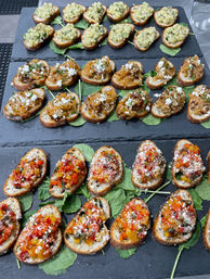 Gourmet appetizer platter of three rows of bruschetta-style crostini on slate boards: herb-and-garlic diced topping, caramelized onions with crumbled feta, and colorful pepper relish with grated cheese, arranged on baby greens — ideal party or catered hors d'oeuvres.