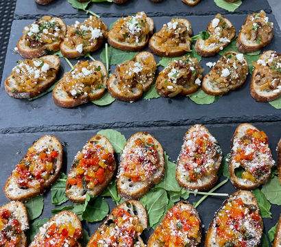 Gourmet appetizer platter of three rows of bruschetta-style crostini on slate boards: herb-and-garlic diced topping, caramelized onions with crumbled feta, and colorful pepper relish with grated cheese, arranged on baby greens — ideal party or catered hors d'oeuvres.