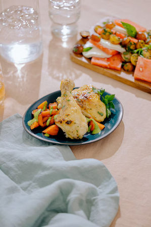 Herb-roasted chicken drumstick and thigh with roasted carrots and green herb sauce on a blue plate, garnished with parsley on a light linen dining table with a charcuterie board in the background