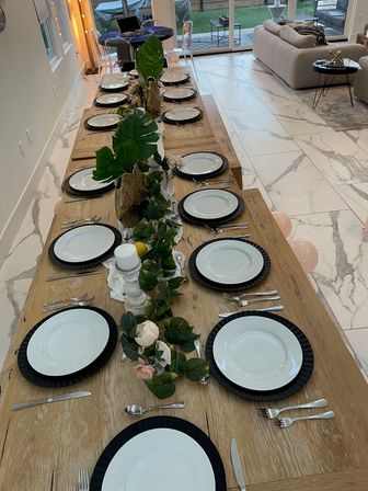 Long wooden dining table set for a dinner party with white plates on black chargers, silver cutlery, leafy floral centerpieces and candles in a modern open-plan living room with marble tile floor and sliding glass doors to a backyard.