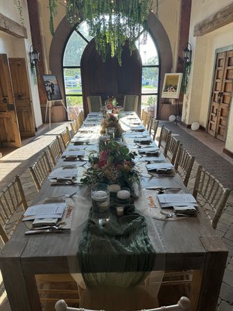 Sunlit rustic barn-style wedding reception table with arched wooden doors, hanging greenery, long wooden banquet table set with floral centerpieces, candles, menus and gold chiavari chairs.