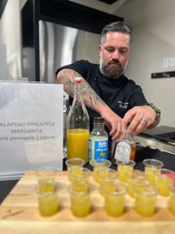 Chef arranging jalapeño pineapple margarita tasting shots on a wooden board, pouring pineapple juice and syrup at a kitchen counter