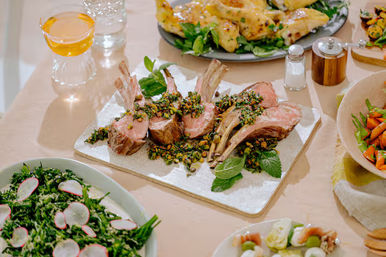 Roasted rack of lamb topped with chopped pistachios and herbs on a white serving board, surrounded by kale-radish salad, roasted carrots and other side dishes on a festive dinner table.