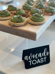 Mini avocado toast appetizers — whipped avocado dollops on round toasted bread, sprinkled with sesame and everything seasoning, arranged on a wooden serving board with a small “avocado toast” sign