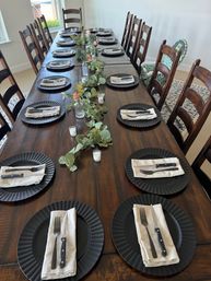Long rustic wooden dining table in a bright dining room set for a large meal with black charger plates, folded linen napkins and knives and forks, eucalyptus garland runner with small vases and votive candles, surrounded by ladder-back wooden chairs and a patterned highchair.