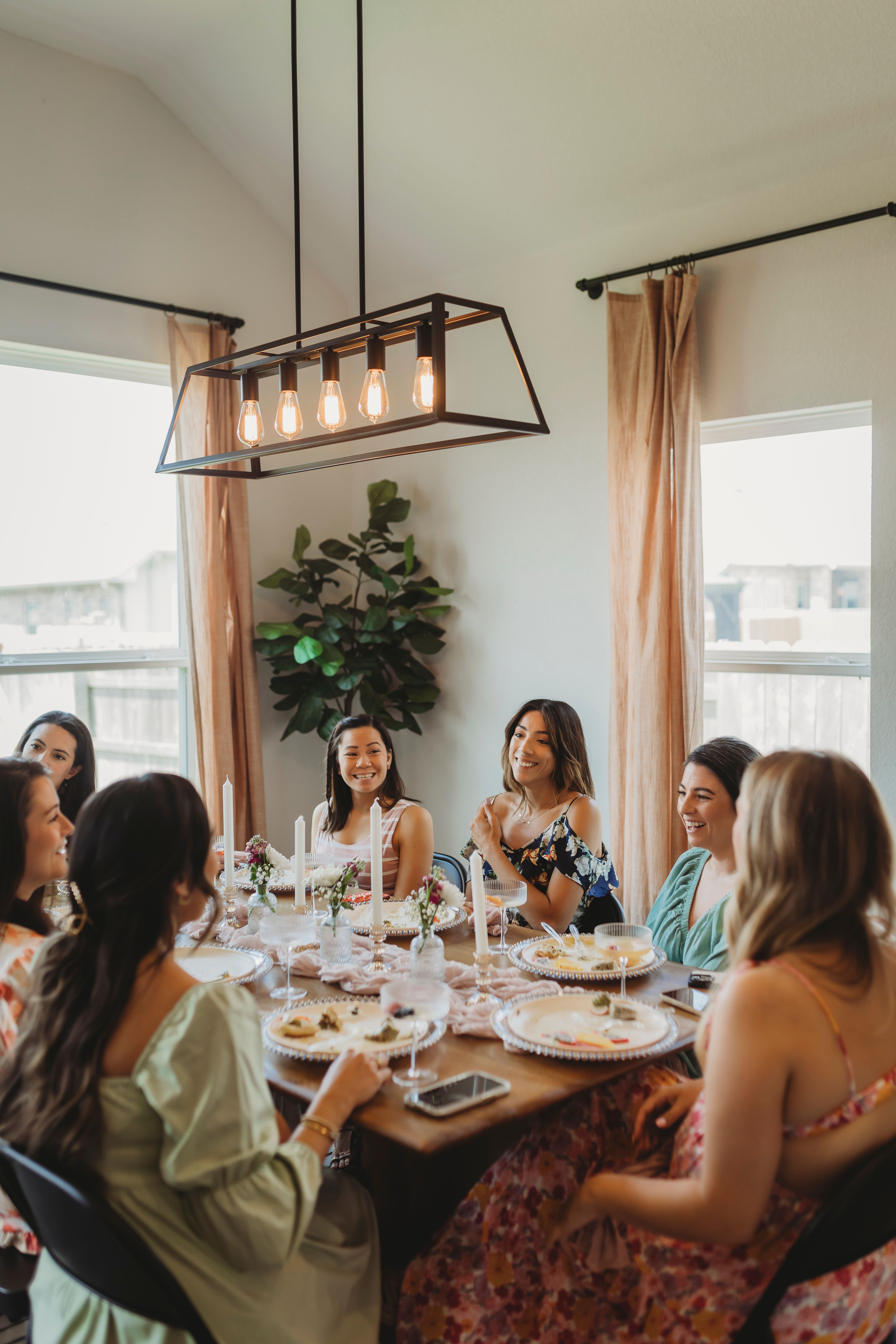 Group of women laughing and enjoying brunch around a decorated dining table in a bright home dining room, featuring a modern rectangular pendant light, tall candles, floral place settings, and beige curtains.