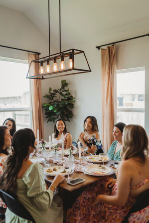 Group of women laughing and enjoying brunch around a decorated dining table in a bright home dining room, featuring a modern rectangular pendant light, tall candles, floral place settings, and beige curtains.