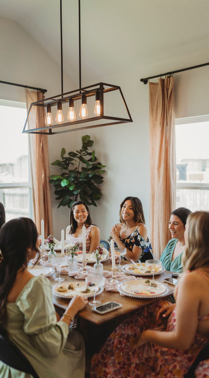 Group of women laughing and enjoying brunch around a decorated dining table in a bright home dining room, featuring a modern rectangular pendant light, tall candles, floral place settings, and beige curtains.