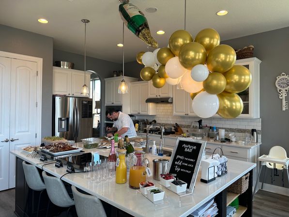 Modern white kitchen with a large island set up for brunch and a mimosa bar — gold and white balloon cluster with a champagne balloon overhead, pitchers of orange juice and mixers, rows of champagne flutes, platters of food, and a person preparing dishes.