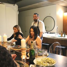 Chef overseeing an intimate candlelit dinner party in a modern home kitchen, guests seated around a table with plated dishes, wine, a champagne bottle and a bowl of pasta.
