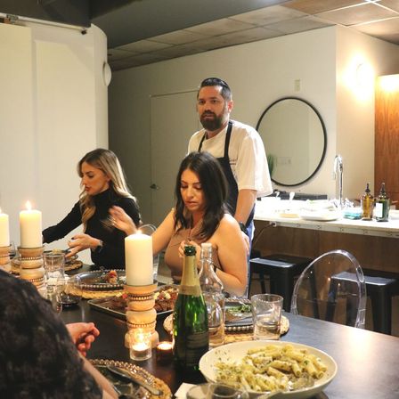 Chef overseeing an intimate candlelit dinner party in a modern home kitchen, guests seated around a table with plated dishes, wine, a champagne bottle and a bowl of pasta.