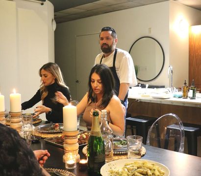 Chef overseeing an intimate candlelit dinner party in a modern home kitchen, guests seated around a table with plated dishes, wine, a champagne bottle and a bowl of pasta.