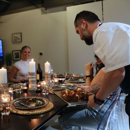 Candlelit indoor dinner party with a server presenting a roasted platter to smiling guests at a modern dining table.