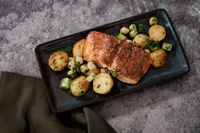 Crispy seared salmon fillet on a black rectangular plate with herb-roasted baby potatoes, diced cucumber and fresh herbs on a gray stone tabletop