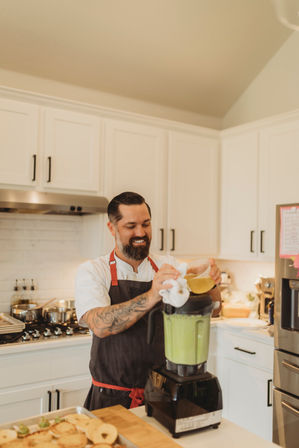Smiling chef in apron pouring dressing into a high-powered blender of green sauce in a bright modern white kitchen during meal prep
