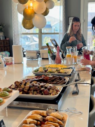 Home brunch buffet on a bright kitchen island — trays of breakfast potatoes, sausages, bacon, scrambled eggs and pastries with juice pitchers, champagne bottles and gold-and-white balloons overhead
