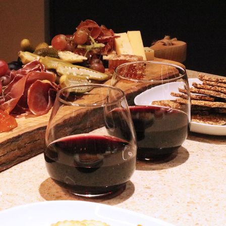 Two stemless glasses of red wine on a countertop beside a wooden charcuterie board with cheeses, cured meats, grapes, pickles and crackers in a cozy indoor setting