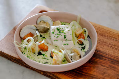 Restaurant-style seafood bowl with a poached white fish fillet, open clams, golden-brown croutons, fennel ribbons and herb cream sauce in a pale bowl on a wooden board