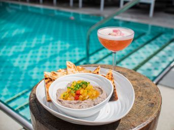 Poolside bowl of creamy bean dip topped with mango salsa and chives, served with toasted pita triangles and a pink floral-garnished cocktail in a coupe glass by a turquoise swimming pool.