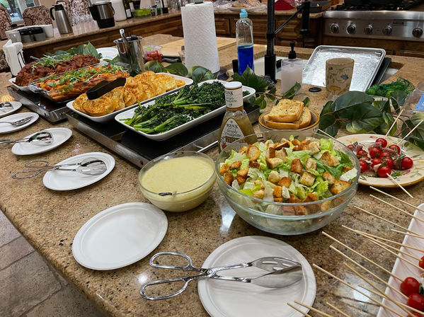 Home kitchen buffet on a granite island: large glass bowl of lettuce and crouton salad with dressing and tongs, skewers of cherry tomatoes, trays of roasted broccolini, seasoned potato rounds, baked pasta and garlic bread, plates and serving utensils.