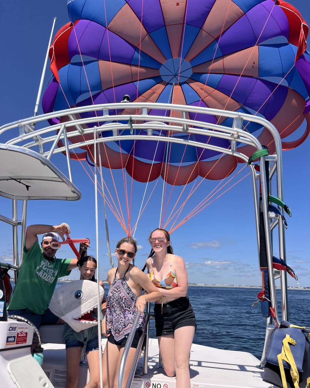 Friends on a parasailing boat off the coast posing on deck beneath a giant purple-blue-red parasail canopy with orange lines over a calm blue ocean on a sunny summer day
