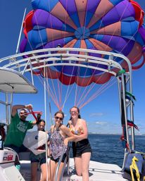 Friends on a parasailing boat off the coast posing on deck beneath a giant purple-blue-red parasail canopy with orange lines over a calm blue ocean on a sunny summer day