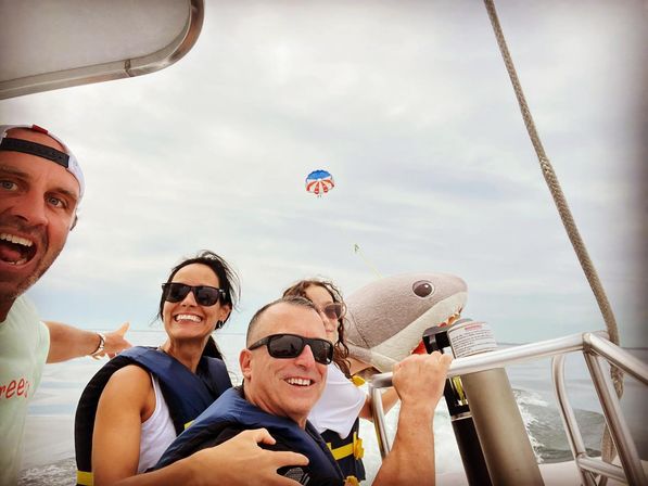 Four adults in life jackets smiling on a speedboat with a large inflatable shark prop and a colorful parasail trailing over the ocean under a cloudy sky.